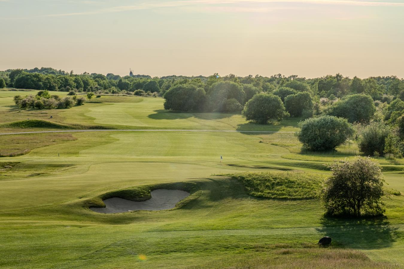 Udsigt over golfbanen ved Hedeland Golfklub udenfor Roskilde med bunker, grønne fairways og Roskilde Domkirke i baggrunden.