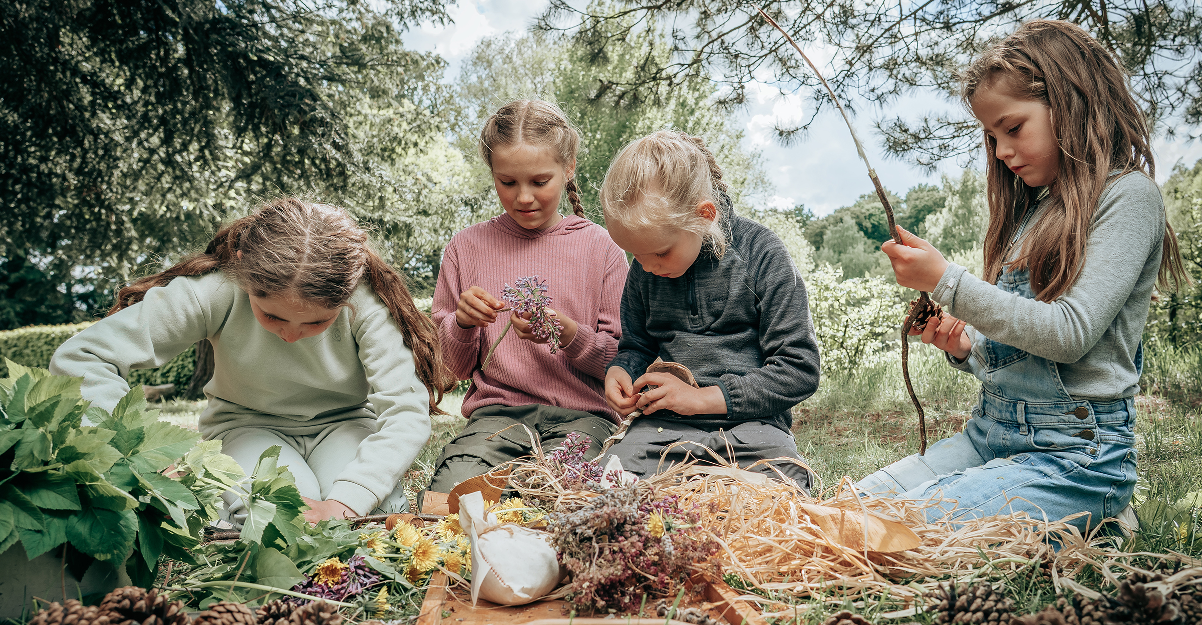 Fire børn sidder i naturen ved Færgegården og laver kreative dekorationer af blomster, blade og naturmaterialer.