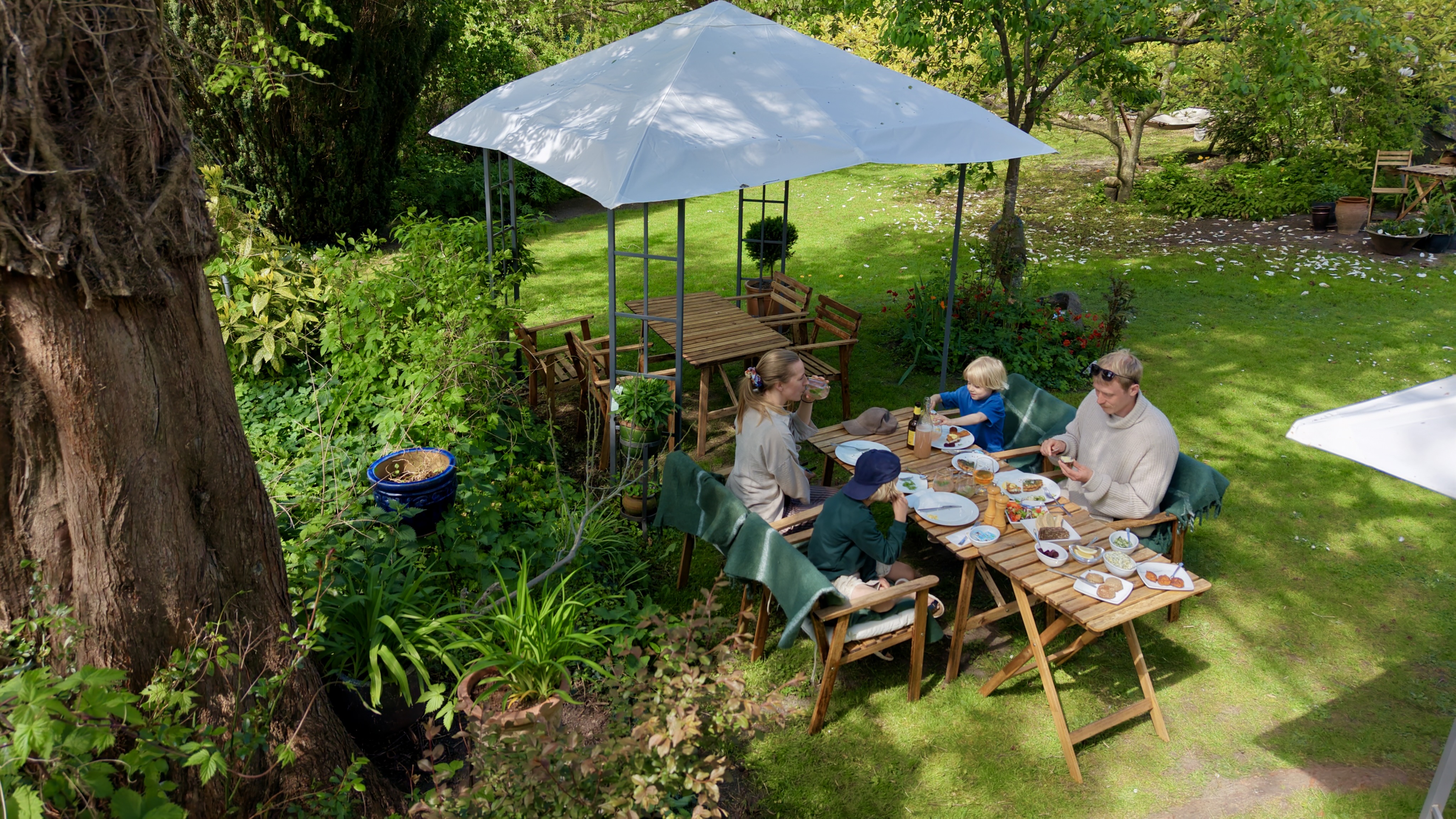 Familie nyder et måltid ved et bord i den frodige have hos Chefs Cottage and Garden Café omgivet af grønne planter og træer.
