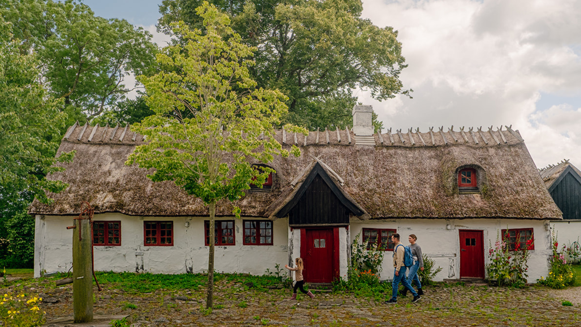 Familie går forbi den historiske stråtækte Gl. Kongsgård i Lejre omgivet af have og gamle træer.