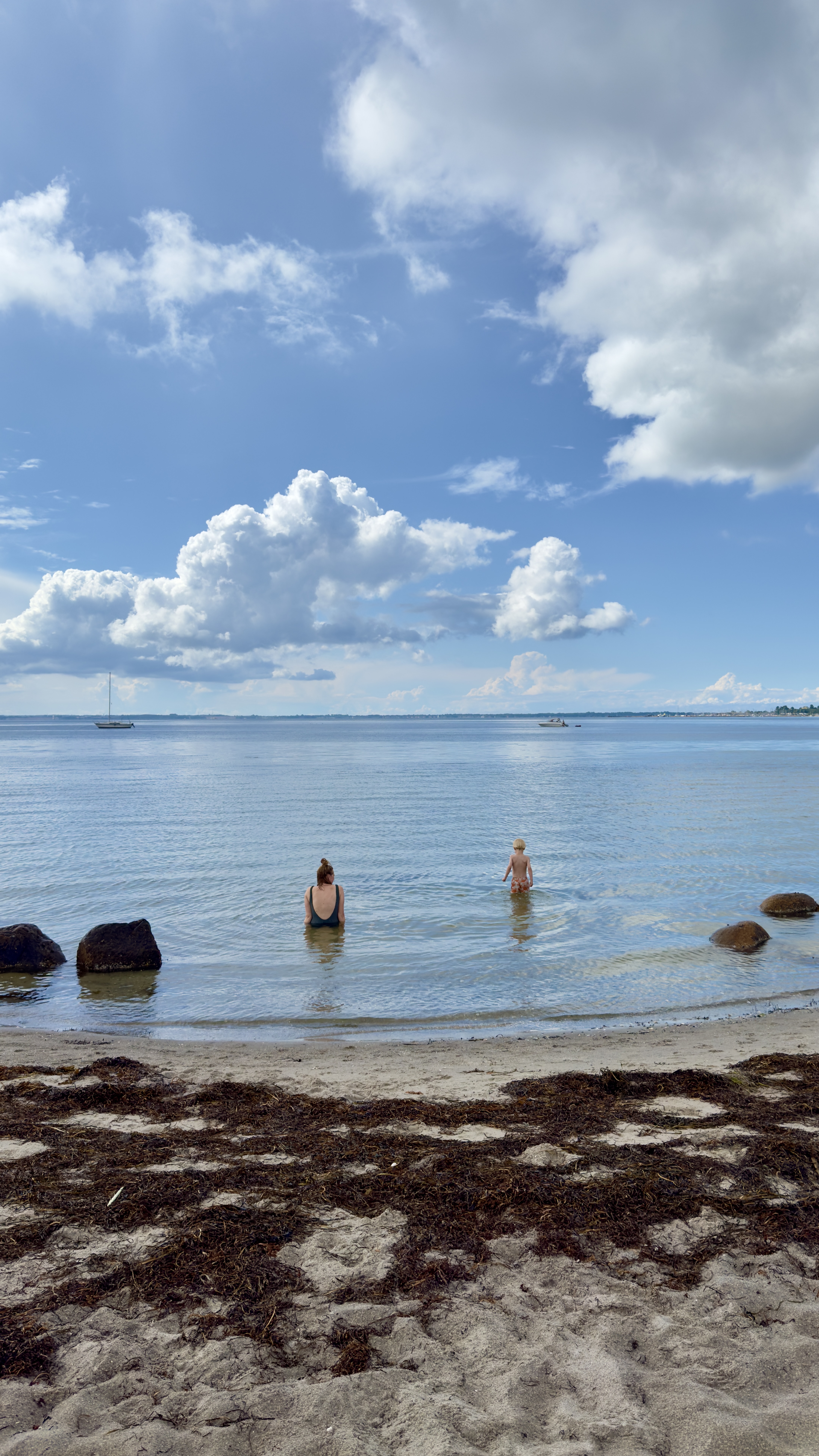 Voksen og barn bader i det rolige vand ved Kulhuse Strand med udsigt over fjorden under en skyfyldt sommerhimmel.