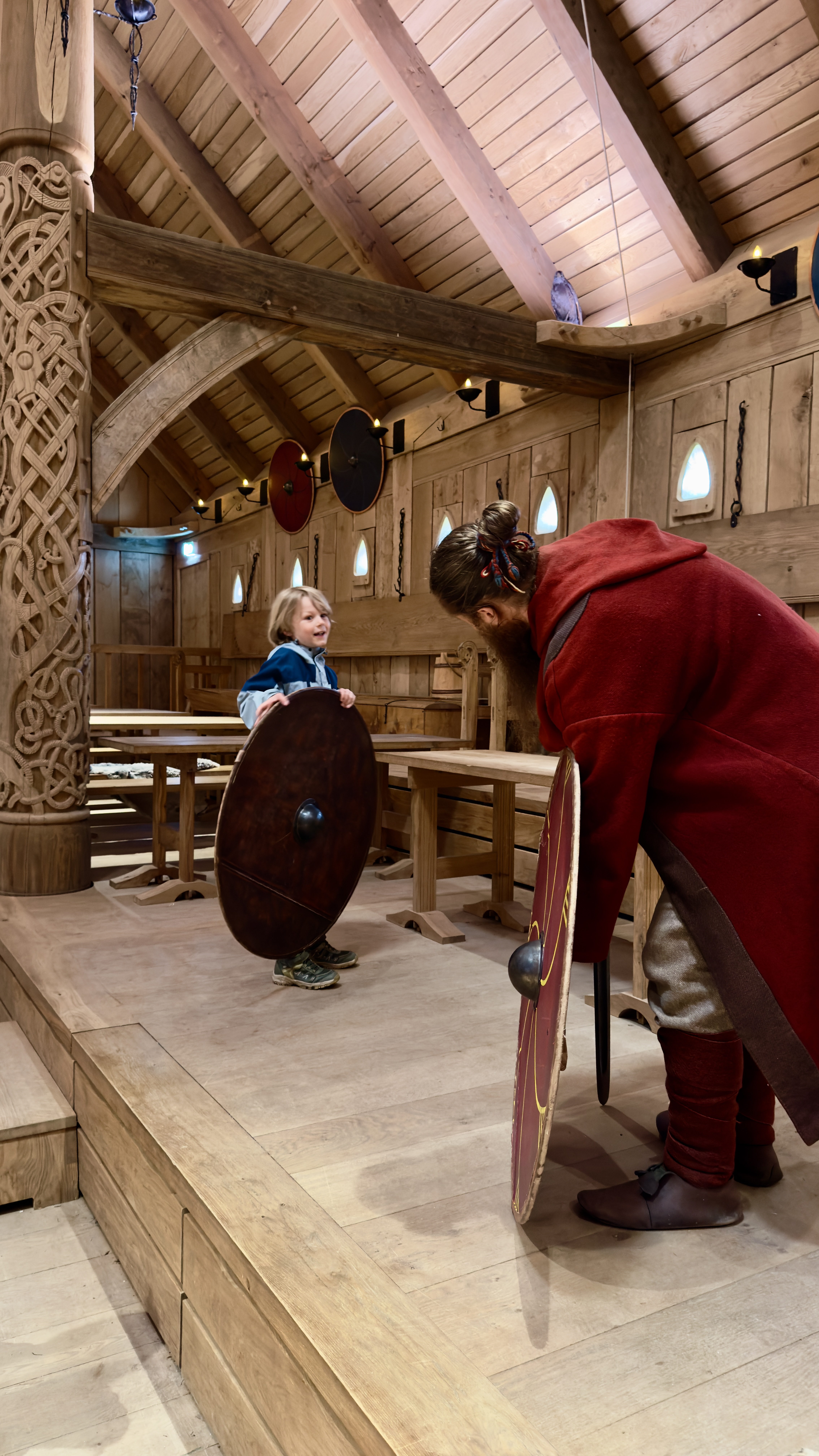 Barn og formidler med skjolde i Kongehallen i Sagnlandet Lejre, hvor besøgende kan opleve vikingetidens kultur og historie.