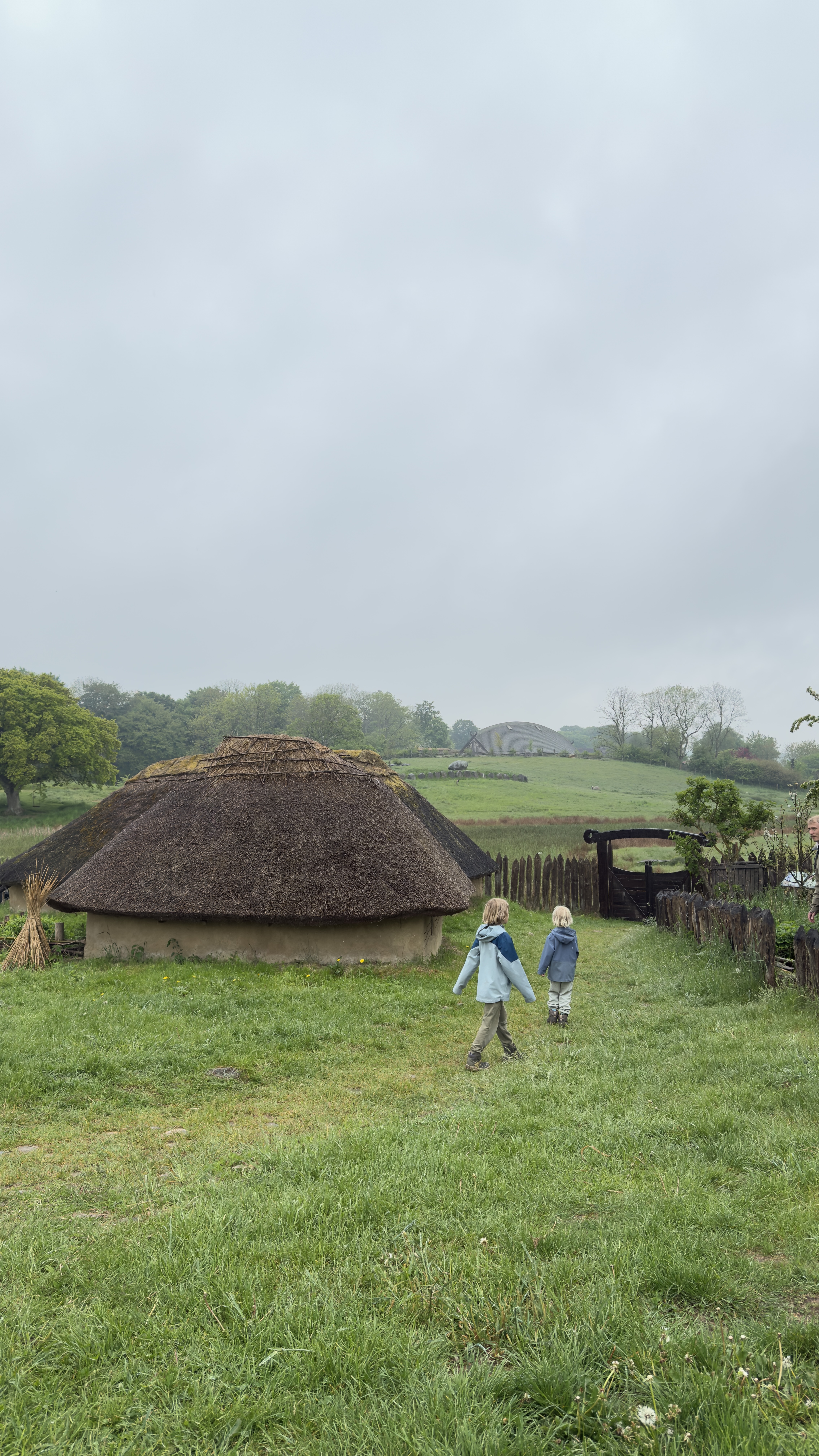 To børn går gennem det historiske jernaldermiljø i Sagnlandet Lejre med stråtækt rundhytte og grønne omgivelser.