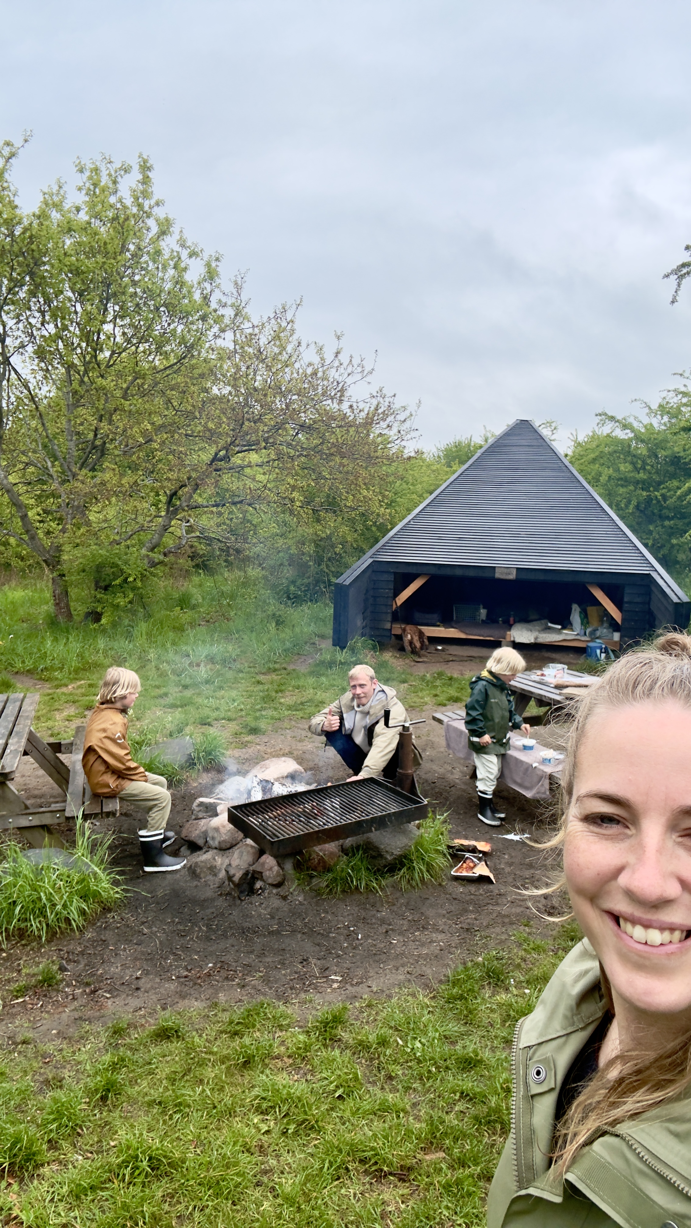 Familie samles omkring bålpladsen ved Lavringe Å Shelterplads med shelter, grill og grønne omgivelser.