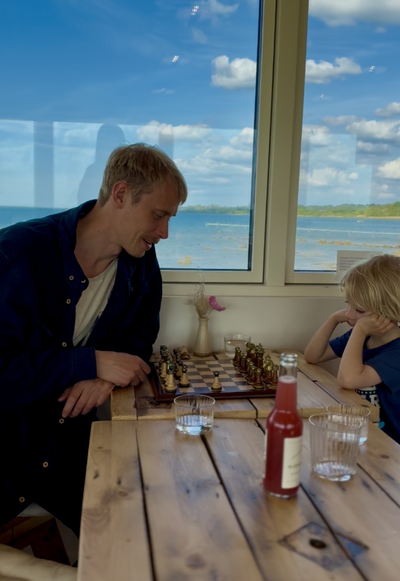 Voksen og barn spiller skak ved et bord med udsigt over fjorden gennem de store vinduer på SeaLevel Kaffebar.