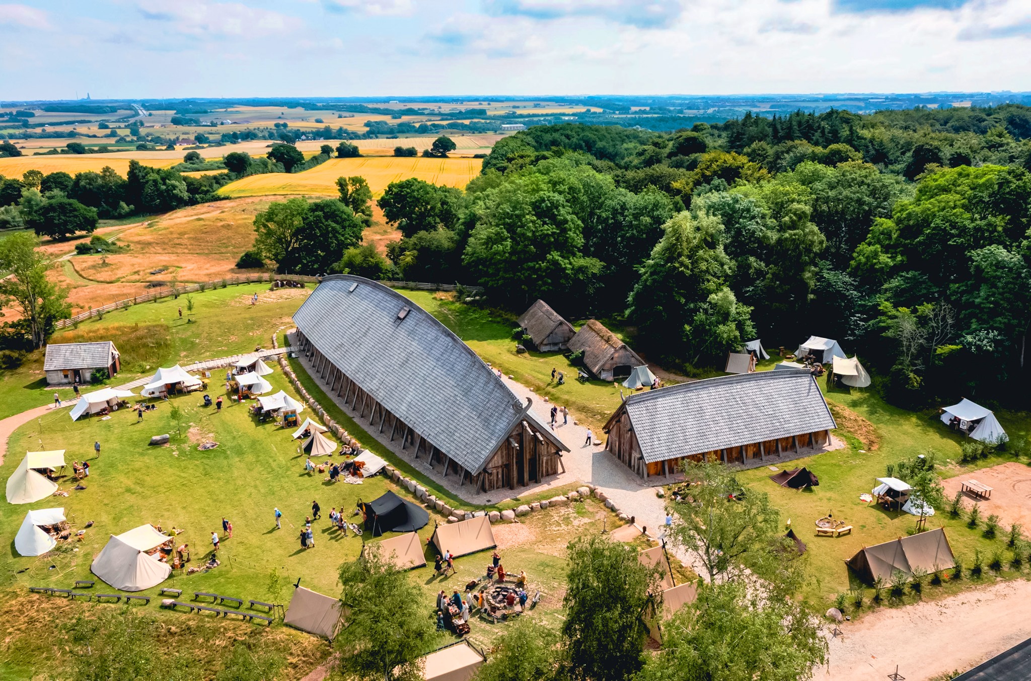 Luftfoto af vikingeboplads med store langhuse, telte og besøgende omgivet af skov og åbent landskab.