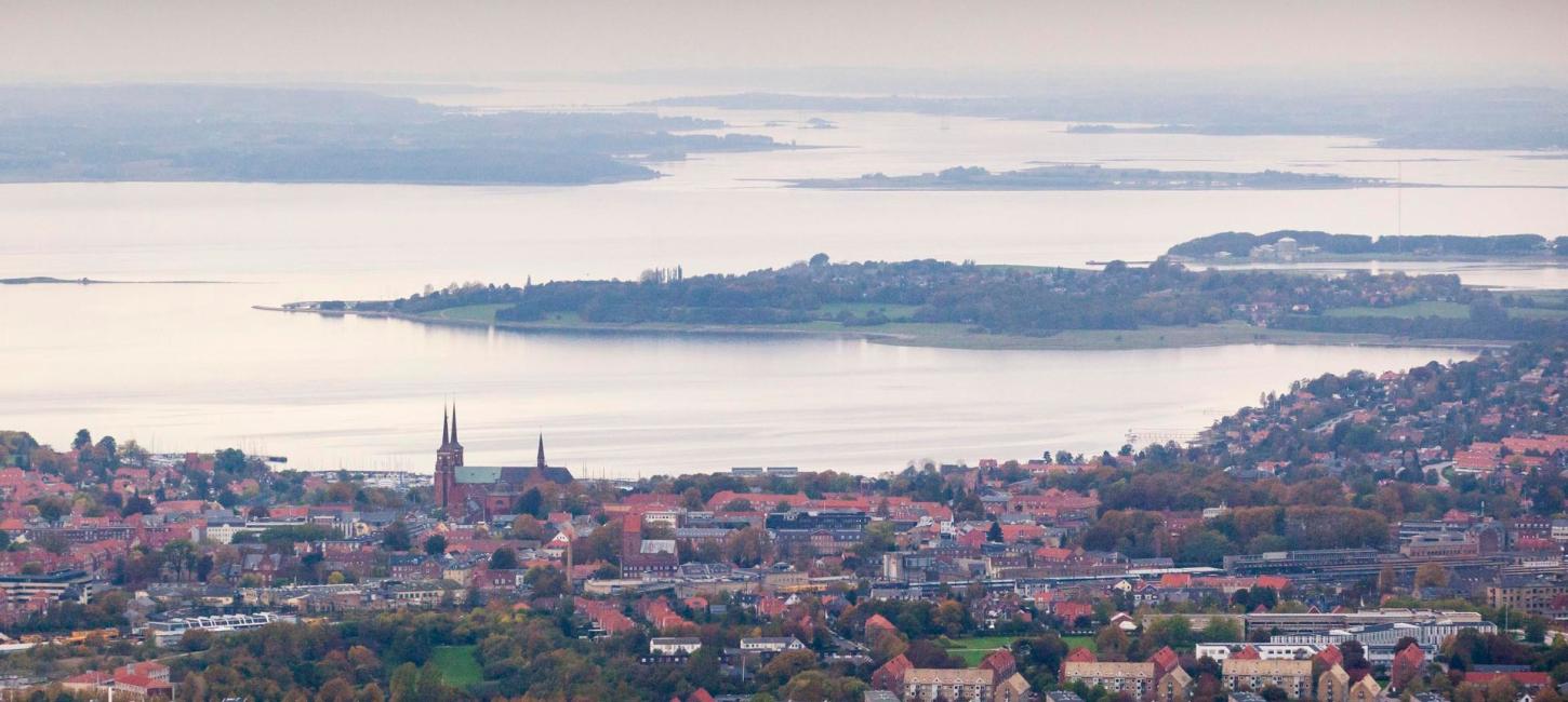 Udsigt over Roskilde med Roskilde Domkirke i forgrunden og Roskilde Fjord omgivet af øer og landskab i baggrunden under en himmel med skyer i aftensol.