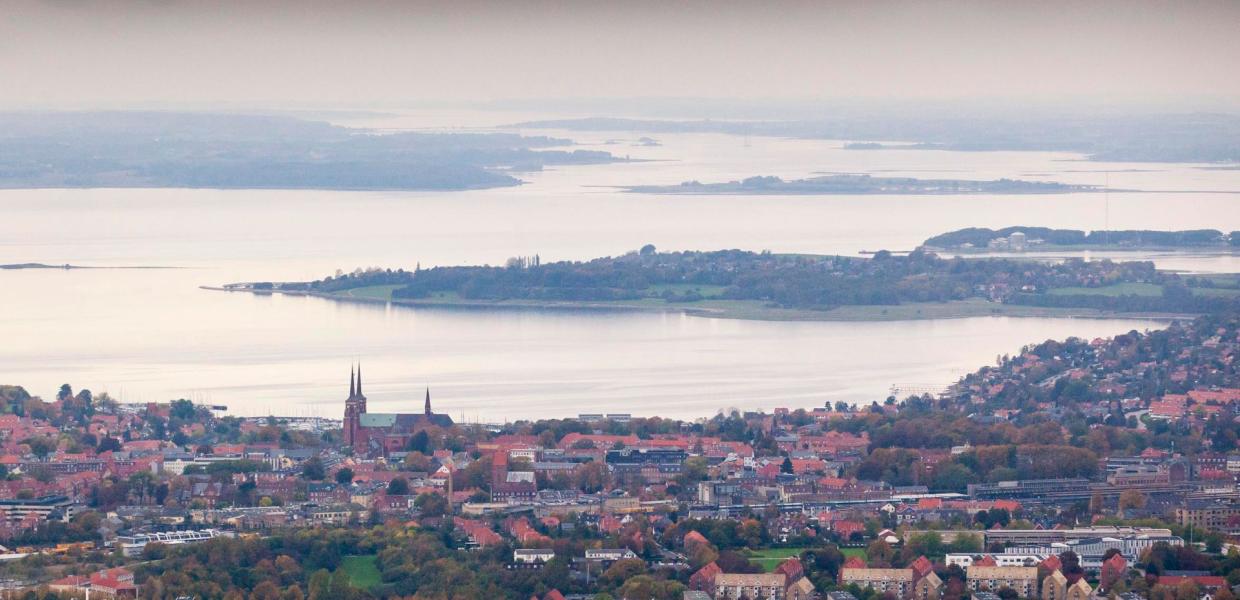 Udsigt over Roskilde med Roskilde Domkirke i forgrunden og Roskilde Fjord omgivet af øer og landskab i baggrunden under en himmel med skyer i aftensol.