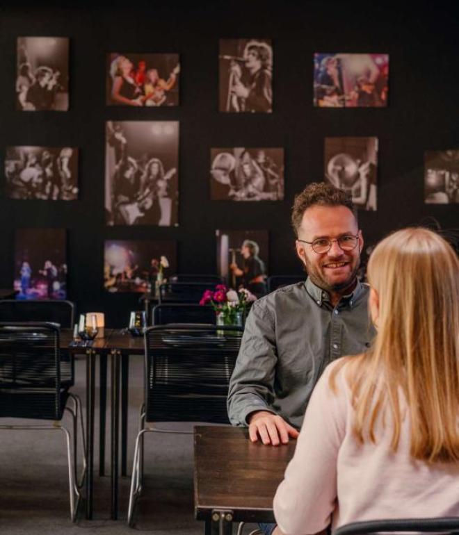 To personer sidder ved et bord i Ragnarocks café, omgivet af vægge dekoreret med rockkoncertbilleder.