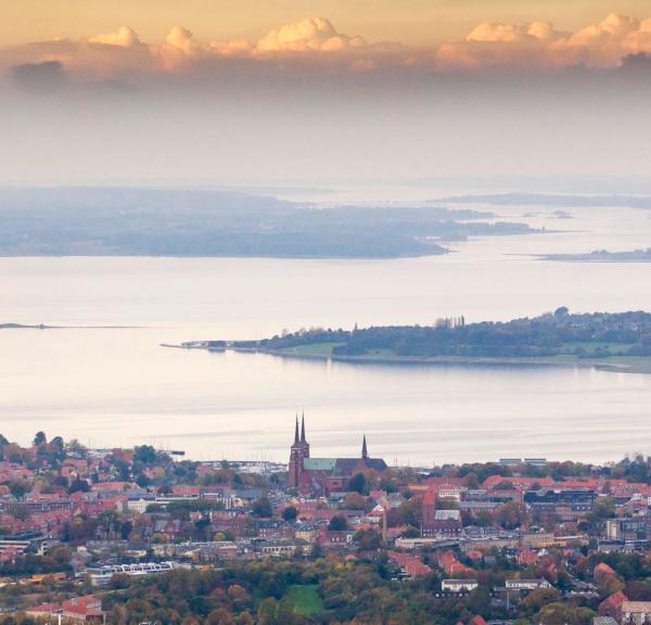 Udsigt over Roskilde med Roskilde Domkirke i forgrunden og Roskilde Fjord omgivet af øer og landskab i baggrunden under en himmel med skyer i aftensol.