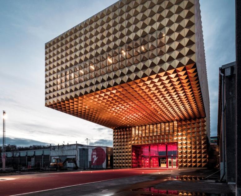 Gylden, kubeformet facade på Ragnarock med markant udhæng og rødlyst indgang i aftenskumring.