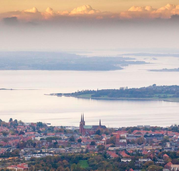 Udsigt over Roskilde med Roskilde Domkirke i forgrunden og Roskilde Fjord omgivet af øer og landskab i baggrunden under en himmel med skyer i aftensol.
